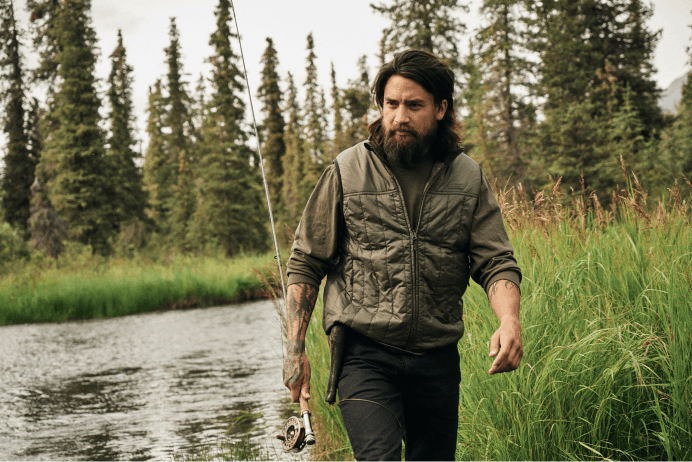 Filson Ultralight Vest in olive grey walking with fishing pole next to river bend in the woods